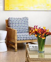 Wicker armchair with blue patterned cushions, flower arrangement, white side table, gold table, yellow painting.