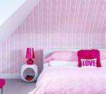 Pink bedroom with slanted wall, patterned wallpaper, bed, lamp, and Love pillow.