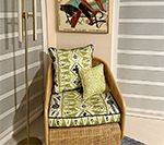 Wicker armchair with green and blue patterned cushions, beneath a framed artwork and gold floor lamp.