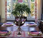 Dining table set for four, centered around a silver centerpiece, with a bright window in the background.
