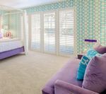 Bedroom with lavender chaise lounge, bed, and white shutters against patterned wallpaper.