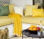 Cozy outdoor seating area with yellow cushions and blanket, green and patterned pillows, and a table with fruit.