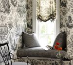 Window seat in a room with toile wallpaper, a small table, a flower vase, and a chair.