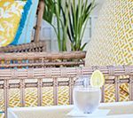 Cocktail with lemon garnish on a table. Wicker chair with yellow and blue patterned pillows.