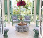 A veranda with teal shutters and furniture, including patterned ottoman, chairs, and stools. Flowers hang above.