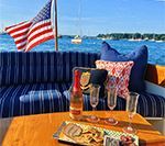 Boat with blue striped cushions, American flag, table with food, drink, and water views.