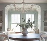 Dining room with round table, arched window, built-in bench, vase of greenery, and chandelier.