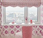 Pink room with window, scalloped shade, chair, and patterned wallpaper, overlooking cityscape.
