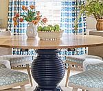 Dining table with blue base, surrounded by patterned chairs, tulips, and a bowl of green apples.