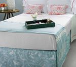 Bed with white bedding, blue blanket and patterned skirt, serving tray with items, side table.