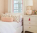 Bedroom with white bed, dresser, pink lamp, and floral pillows.