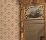Gold-framed mirror with a painted ship scene, reflecting a patterned wall.