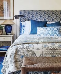 Bedroom with blue patterned headboard, bedding, and accent pillows. White nightstand with blue lamp.