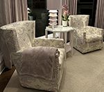 Two upholstered chairs, small table, and decorative tower in a room with a rug and curtains.