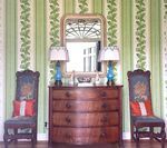 Chest of drawers with mirror, lamps, and chairs against green patterned wallpaper.