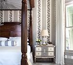 Bedroom with wooden four-poster bed, patterned wallpaper, nightstand with lamp, and window with natural light.