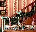 Christmas decorations: garland, bows, gingerbread houses, trees on a staircase and dresser, with patterned wallpaper.
