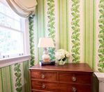 Chest of drawers with lamp and flowers next to window with green leaf wallpaper.