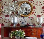 A wood buffet with a mirror and floral arrangement, against patterned wallpaper.