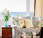 Living room with ocean view: gray sofa, floral pillows, turquoise vase with lilies, wooden side table.
