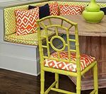 Yellow bamboo-style chair with patterned seat cushion, near a built-in corner bench with orange and yellow pillows.