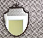 Shield-shaped mirror with dark frame, decorative top detail. Hanging on a patterned wall, reflecting a light-colored interior.