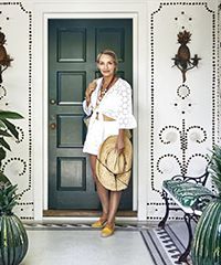 Woman in white outfit stands by green door, holding a straw hat.  Decorative facade with pineapple sconces.