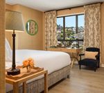 Cozy hotel room with bed, lamp, desk, chair, and patterned curtains framing a city view.