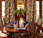 Dining room with round wood table set for dinner. Floral patterned curtains and ornate chairs.