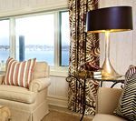 Cozy living room with cream armchair, striped pillow, view of water through window, and patterned curtains.