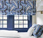 Blue and white patterned Roman shade above a blue framed window. White bedding and throw.