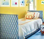 Yellow-walled child's bedroom with upholstered bed in blue geometric print. Artwork on wall, white bedding with orange trim.