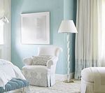 A light blue bedroom with a white armchair, floor lamp, and artwork.