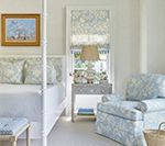 Bedroom with light blue floral accents. White bed, chair, walls, and side table. Small painting on wall.
