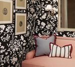 Pink sofa with grey and striped pillows in front of black and white floral wallpaper, framed art.