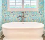 Oval bathtub with chrome faucet against blue floral wallpaper, under a window.