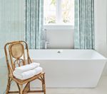 Bathroom with white bathtub, rattan chair holding towels, and blue striped curtains.
