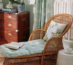 Wicker chaise lounge with blue cushion and pillow, next to a small table. Brown dresser and window visible.