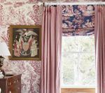 Room with patterned wallpaper, framed artwork, pink curtains, and a Roman shade over a window.