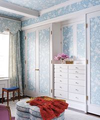 Blue floral-wallpapered closet interior with white trim, drawers, and an ottoman with a red dress on top.