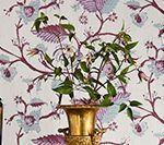 Golden vase with green foliage and white flowers against a floral wallpaper backdrop in purple and blue.