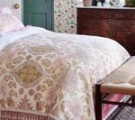 Bed with ornate cream and gold comforter, wood dresser, floral wallpaper.