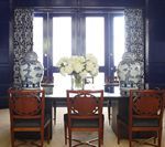 Dark blue room with a table, chairs, and decorative jars flanking the window. White flowers in a vase on the table.