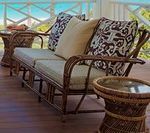 Rattan sofa with cream cushions and patterned pillows on a wooden deck, two matching side tables.
