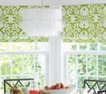 Two windows with green patterned Roman shades, white chandelier.