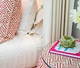 Close-up of a white upholstered chair with red and white patterned pillows, a patterned side table with a book, and white curtains.