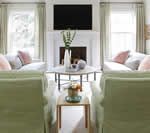 Living room with green armchairs facing each other; pink pillows on light gray sofas, white fireplace, TV above.