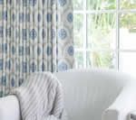 White couch with a striped blanket; patterned blue and white curtains, next to a window.