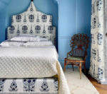 Blue bedroom with patterned headboard, bedspread, and curtains; wicker chair on rug.