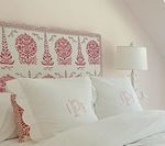 Bed with white linens, pink embroidered headboard and pillows, lamp on nightstand.